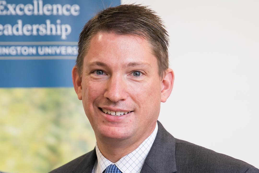 A man smiley brightly in a grey blazer while standing beside a banner of George Washington reading "Center for Excellence in Public Leadership". 