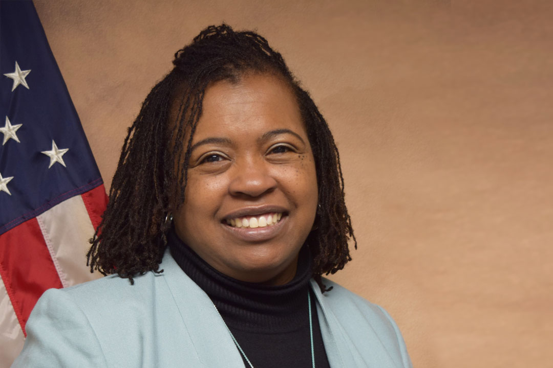 Headshot of a woman with the American flag in the background and wearing a light gray jacket, dark shirt. She has shoulder length hair and a broad smile.