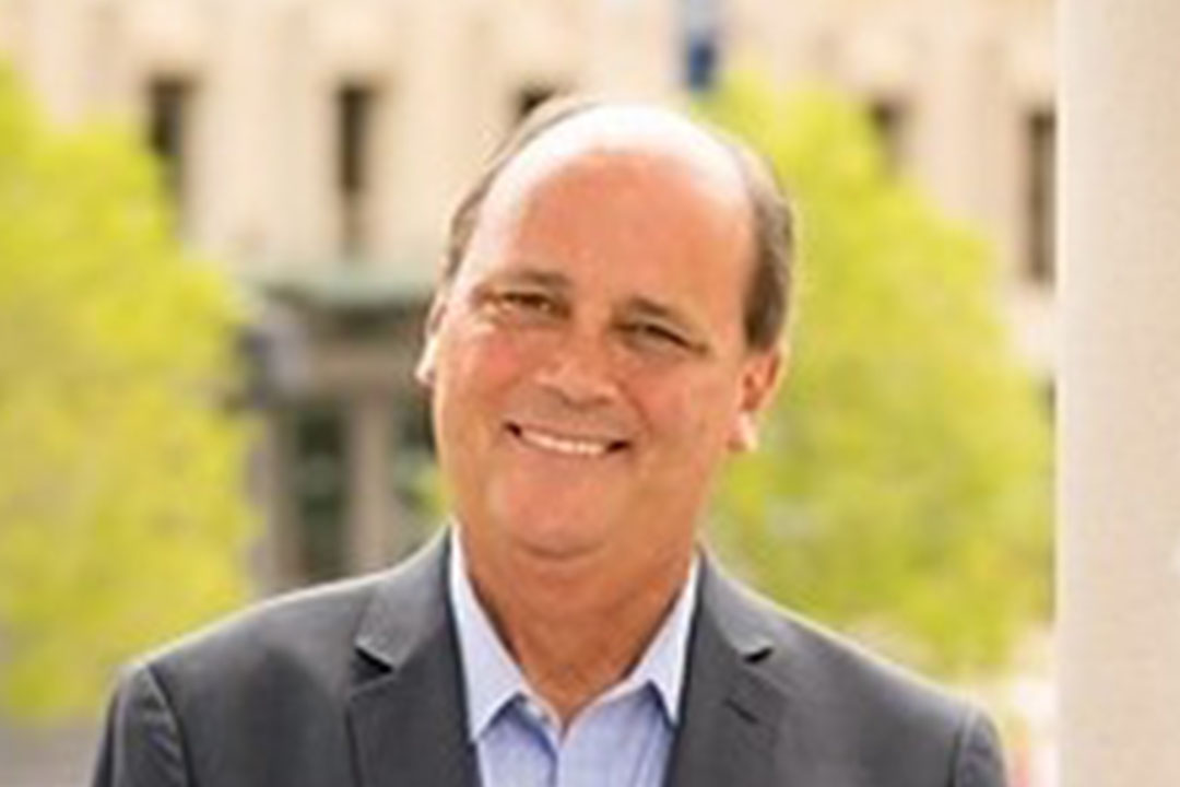 Headshot of man with gray suit and light blue shirt smiling broadly, a building with tall shrubs is behind him in the distance