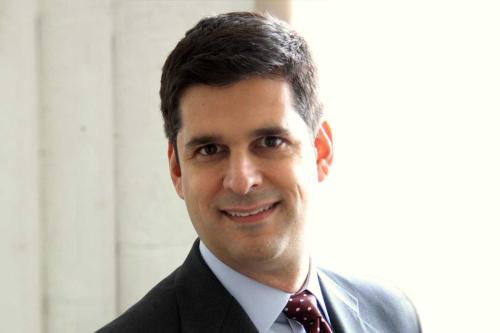 Man in a dark suit with a light blue shirt smiling in front of a bright window