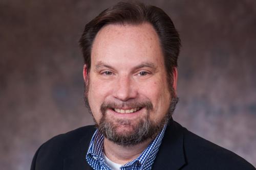 Man in a dark jacket with a blue and white checked shirt smiling broadly
