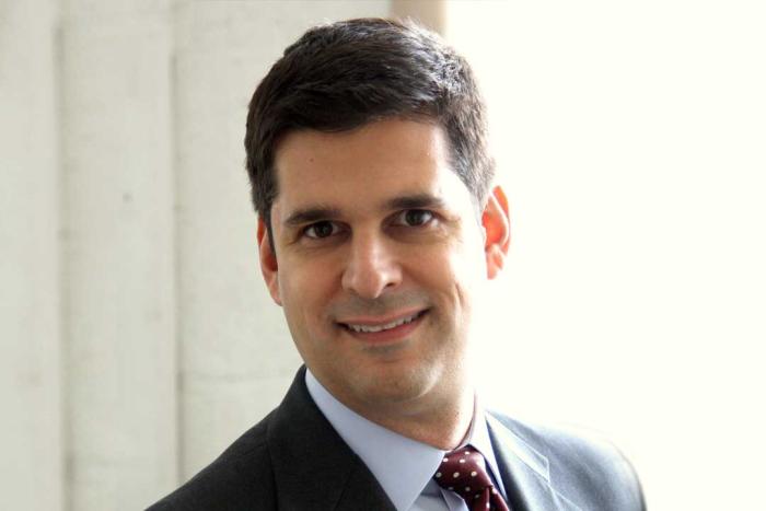 Man in a dark suit with a light blue shirt smiling in front of a bright window