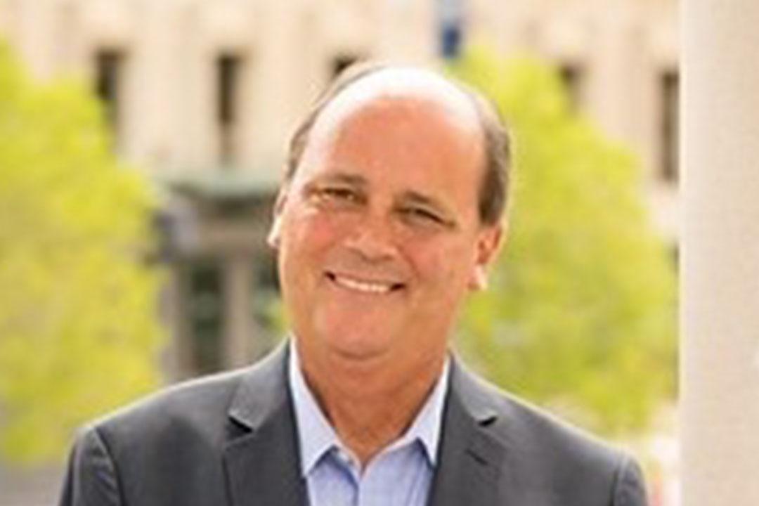 Headshot of man with gray suit and light blue shirt smiling broadly, a building with tall shrubs is behind him in the distance